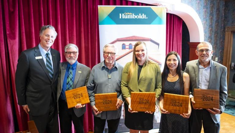 Five smiling alumni award winners with their awards posing with the Cal Poly Humboldt President.