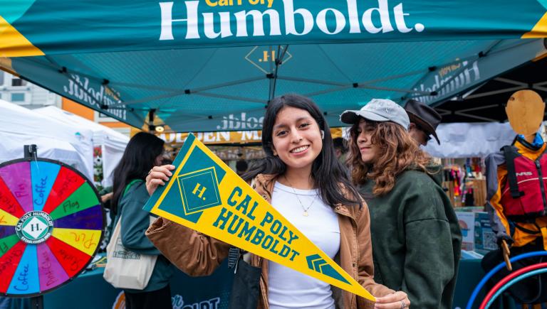 Student with pennant in front of the Cal Poly Humboldt Tent at Farmers' Market