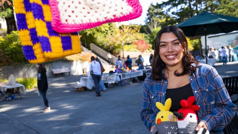 A student with crochet animals at the Makers Market with sales tables in the background.