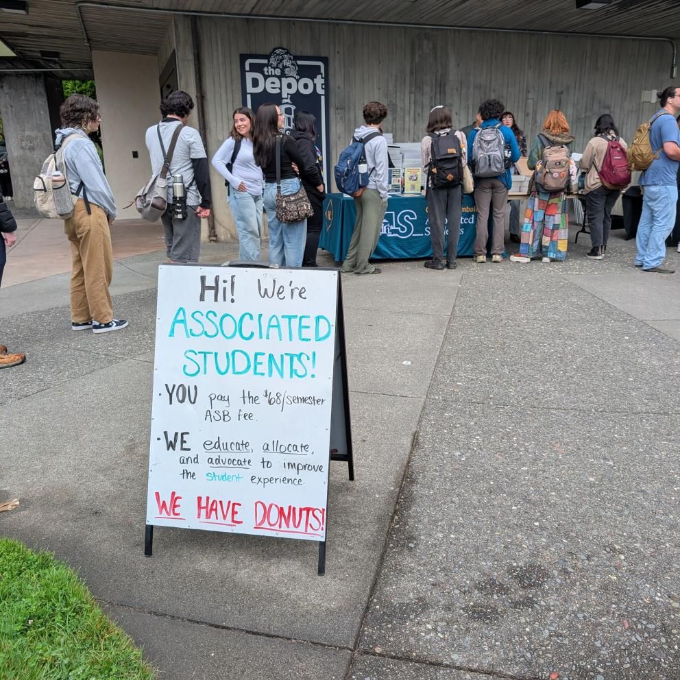Students standing in line to receive donuts. Sign says: Hi! We're Associated Students! You: pay $68/semester student fee. We: Educate, Allocate, and Advocate on behalf of the student population. We have donuts!