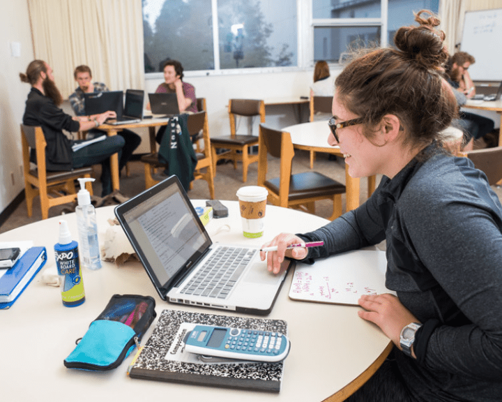 Student working on a laptop and whiteboard at a study table, with others collaborating in the background.