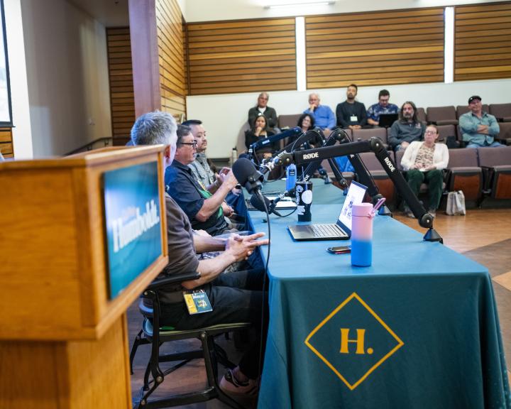 Panelists sitting at table next to podium with Rhode mics. People in audience and in background. Whiteboard with presentation behind them.