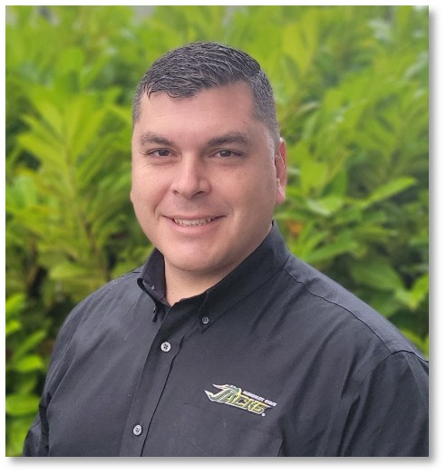 photo is of a man from the shoulders up with green bushes in the background. the man has short dark hair and is wearing a black button up shirt. the man is smiling at the camera.