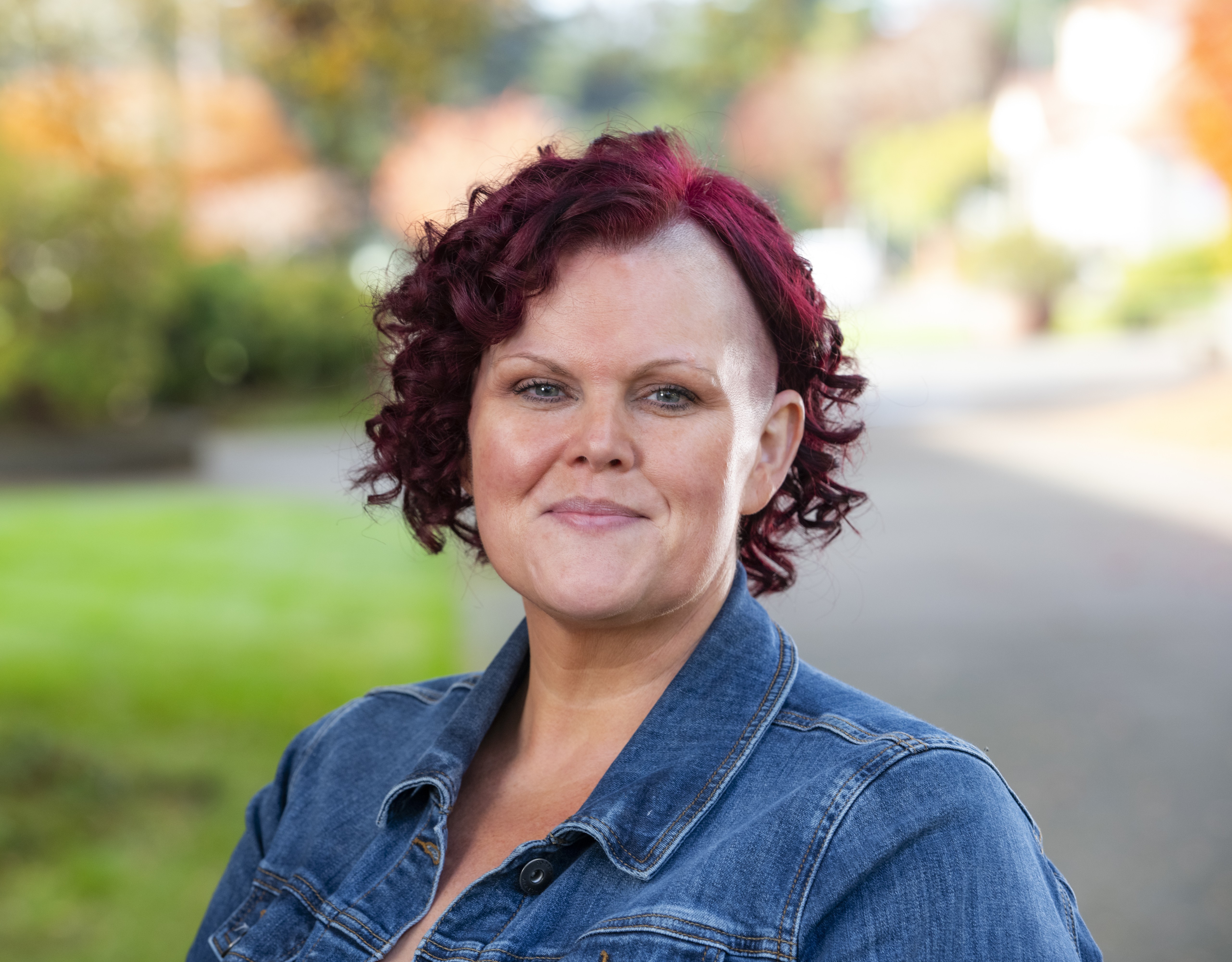 A woman with dark red hair, cropped on one side, she's wearing a denim shirt, and standing in front of a lawn and road.