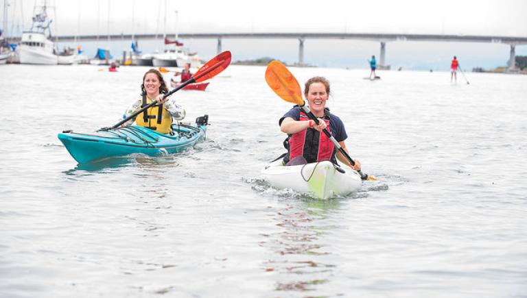 Two people paddling in kayaks on the Humboldt Bay with a large bridge in the background.
