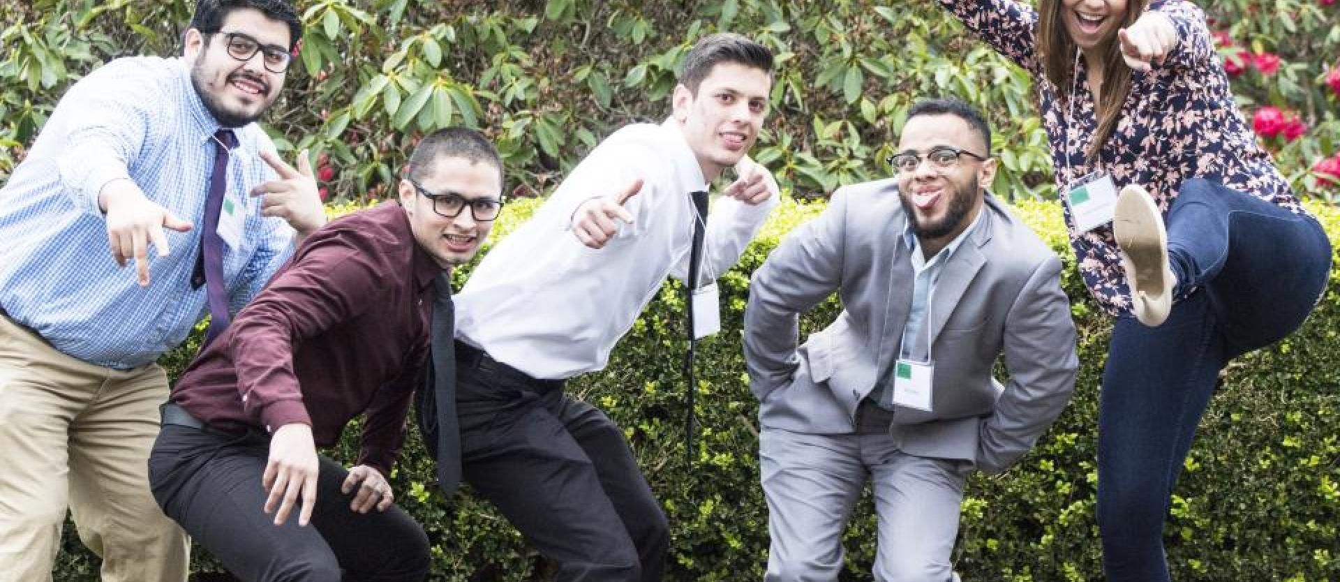 five students crouching in front of a bush, smiling and pointing at the camera 