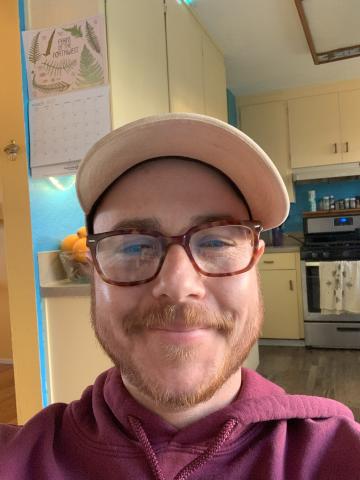 Elias is smiling wearing brown glasses, a tan hat, and a maroon sweater.