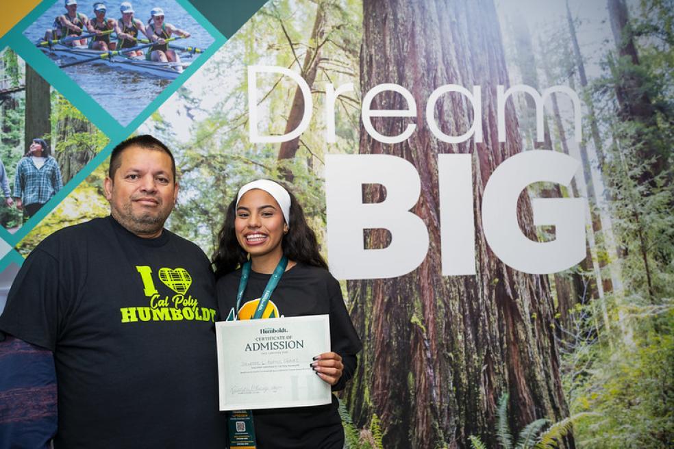 student and familymember stand in front of a sign that reads "Think Big" while holding an acceptance letter to Humboldt