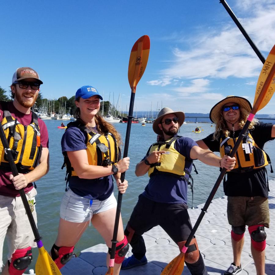 Four students in life jackets standing on the dock in front of Humboldt Bay holding kayak paddles