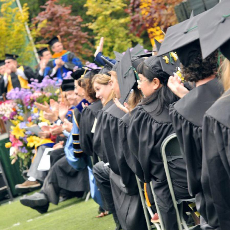 Looking over the shoulder of a row of students in black caps and gowns at graduation. In front of them is a stage with several others in robes seated on it