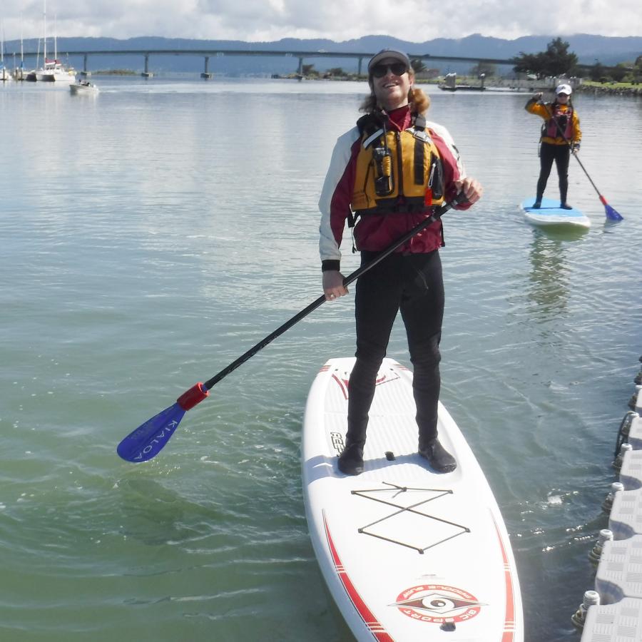 An individual in a wetsuit, red and white splash top, sunglasses, hat, and yellow personal flotation device stands on a paddle board, holding a paddle. He is in a bay with another paddleboarder behind him in the background. 