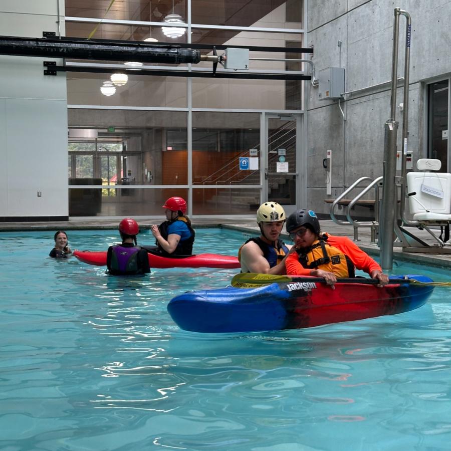 A person in a blue a red river style kayak is being supported by another person in a shallow pool, learning to roll the boat. They are wearing helmets and protective gear. There is another kayaker in a red boat behind them with another instructor.