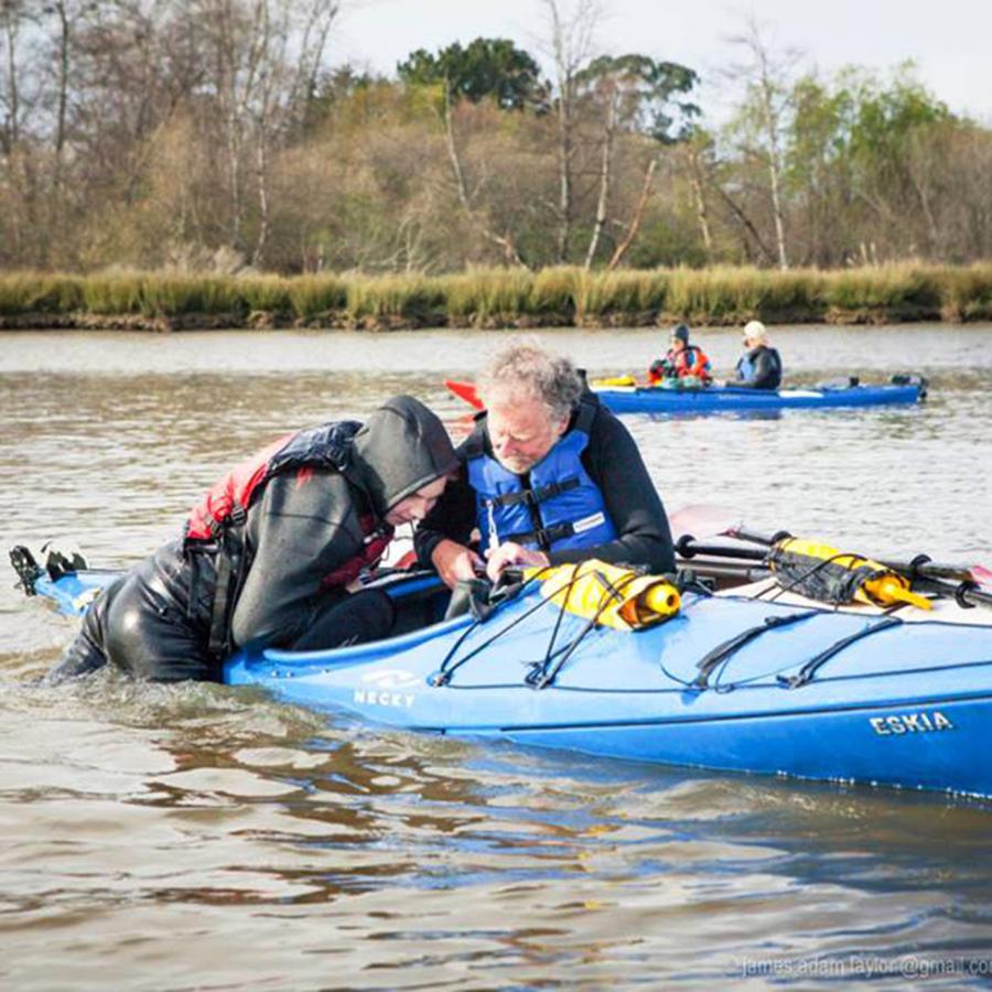 A kayaker dressed in black is pulling themselves back into a blue sea kayak. Another participant is holding their boat next to them. There are two kayakers in the background along with trees and bushes