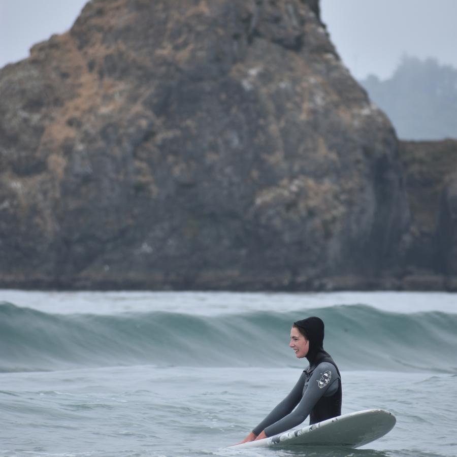 A surfer in a wetsuit with a full hood stands in waist deep water with their surfboard. A wave approaches and a large rock stands in the background.