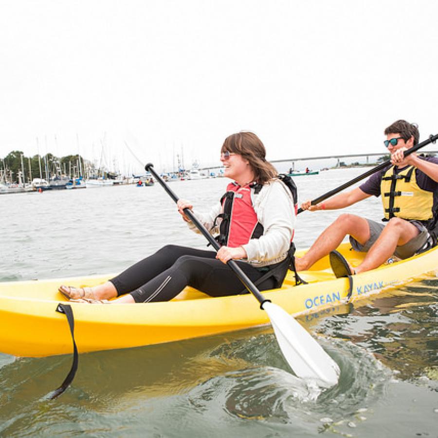 A yellow double kayak on the water with two people sitting in it. They are both wearing life jackets and holding paddles