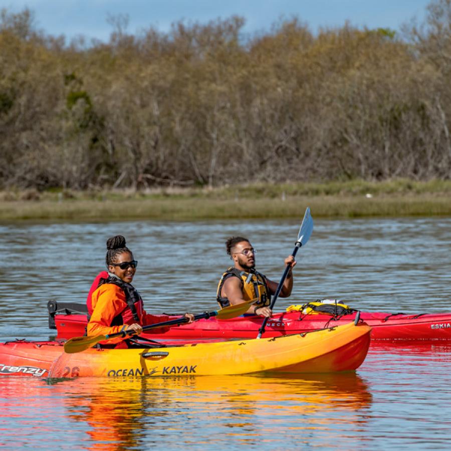 Two kayakers in boats holding paddles and wearing life vests. One kayak is orange and the other is red. They are in the water and in the background there is shoreline and trees and bushes