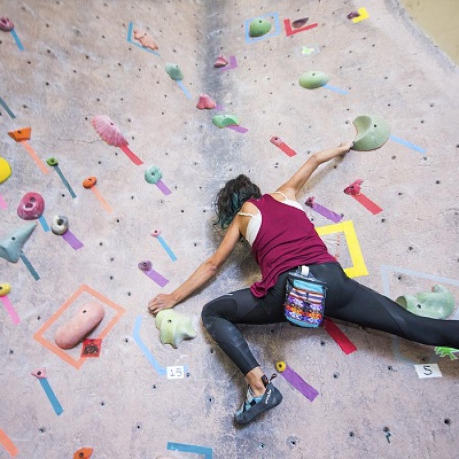 A indoor rock climber positioned on an artificial rock wall. There are handholds of many different colors with pieces of tape indicating their position. The climber is outstretched and reaching for a far away hold