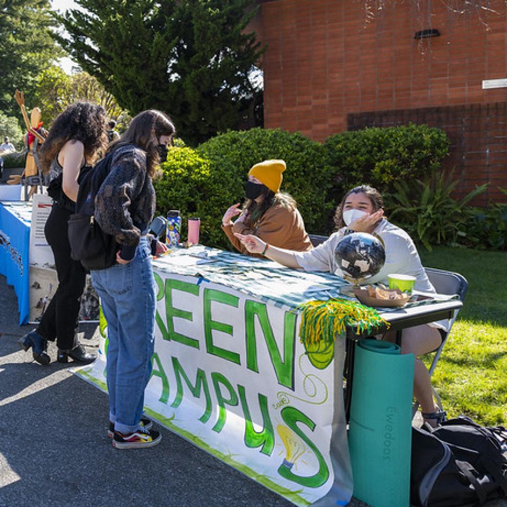 students wearing masks sitting behind a table with a green campus banner on it
