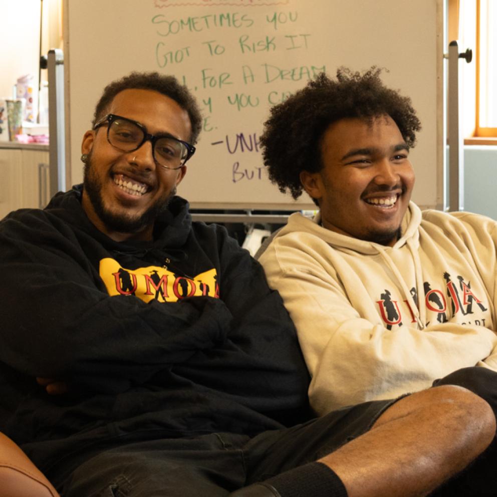Two Umoja Students pose and smile in the Umoja Center wearing Umoja branded hoodies, in colors tan and black.