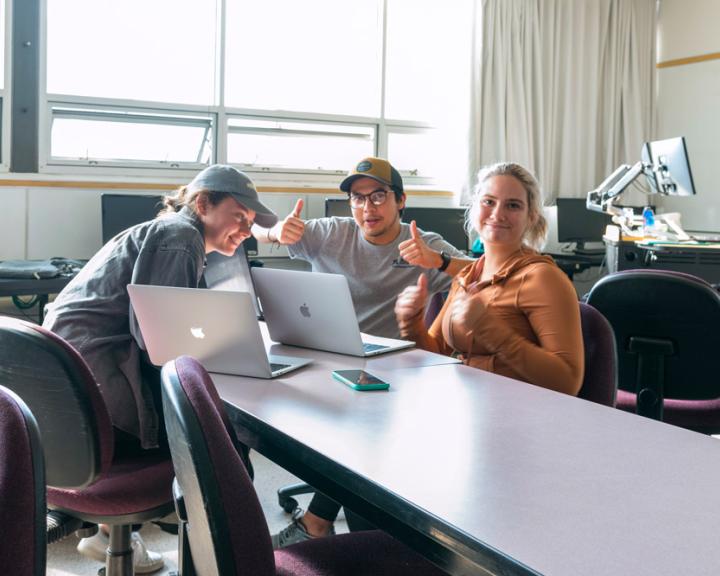 students sitting at a table with computers smiling at the camera