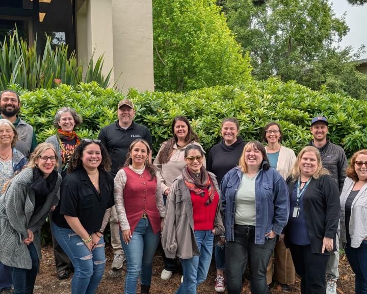 Group of Academic Advising Center Advisors smile while standing in front of pretty landscaping on campus..