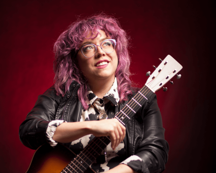 A young white woman with long purple hair and glasses holding a guitar