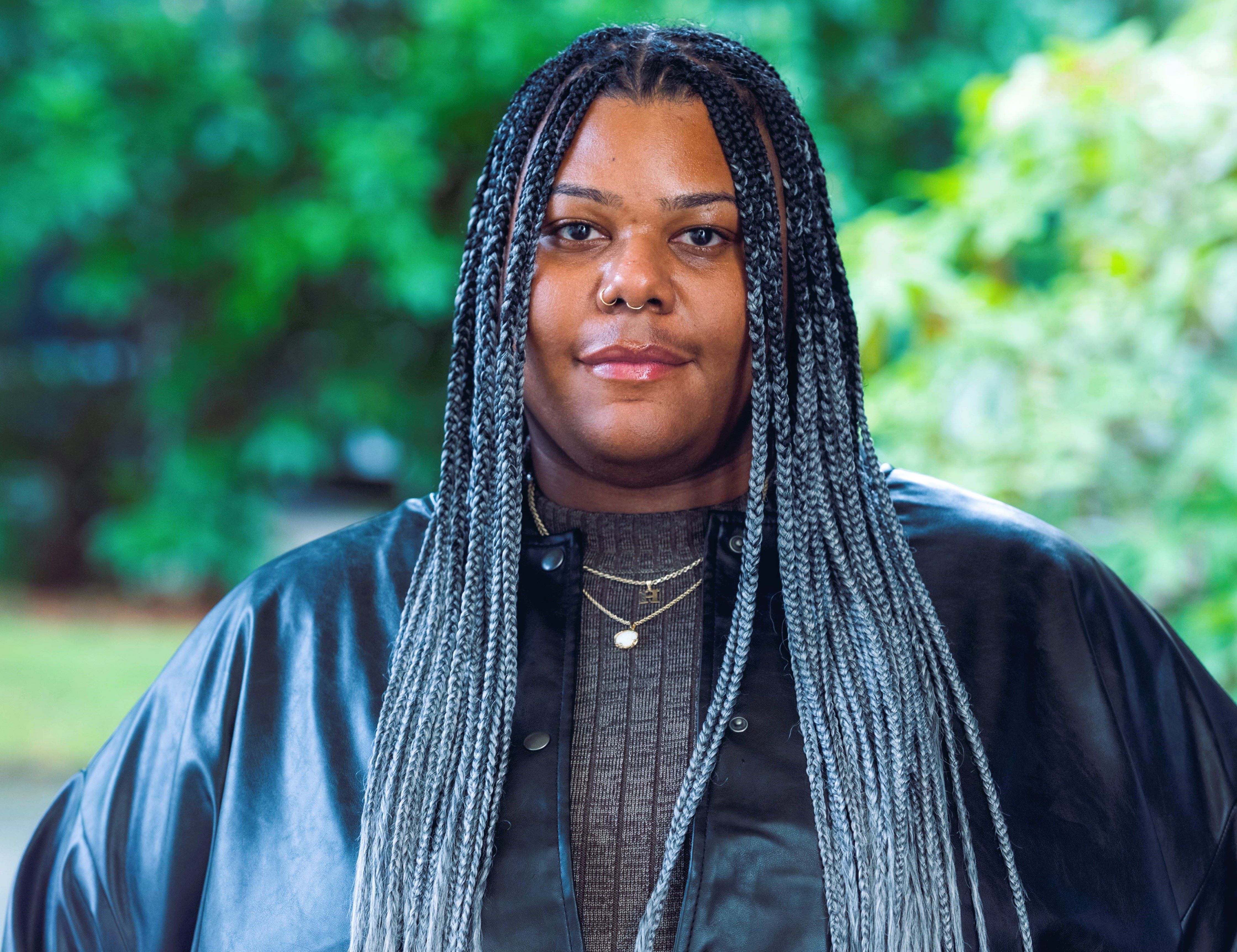 Alexus-Harrelle Deshazier, the coordinator for the Umoja Center. She is smiling wearing an olive/brown mock neck sweater with leather jacket. Her hair is in the style of salt and pepper knotless braids.